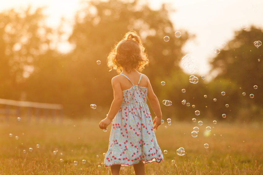 Young child standing in a sunlit field surrounded by floating bubbles