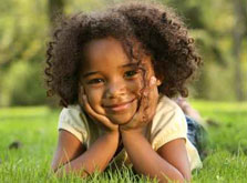 A smiling female child laying on grass with her head resting in her hands